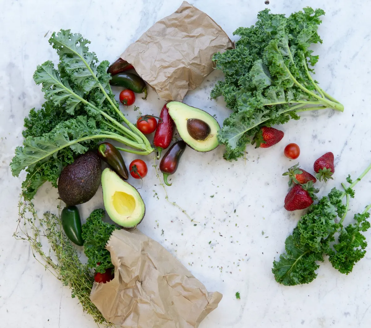 An overhead spread of fresh fruits and vegetables: avocados, tomatoes, peppers, citrus, and leafy greens on a pale surface.