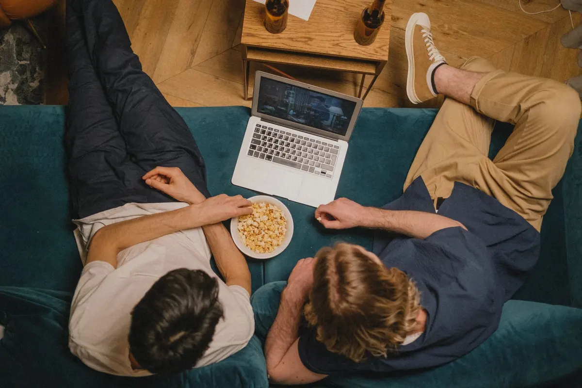 Two people on a couch watching a show on a laptop with a bowl of popcorn between them.